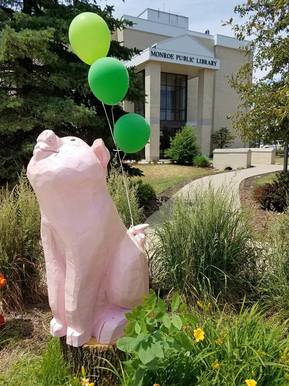 Wooden sculpture of Wilbur the Pig with balloons on his tail, with the library building in the background, summer 2017